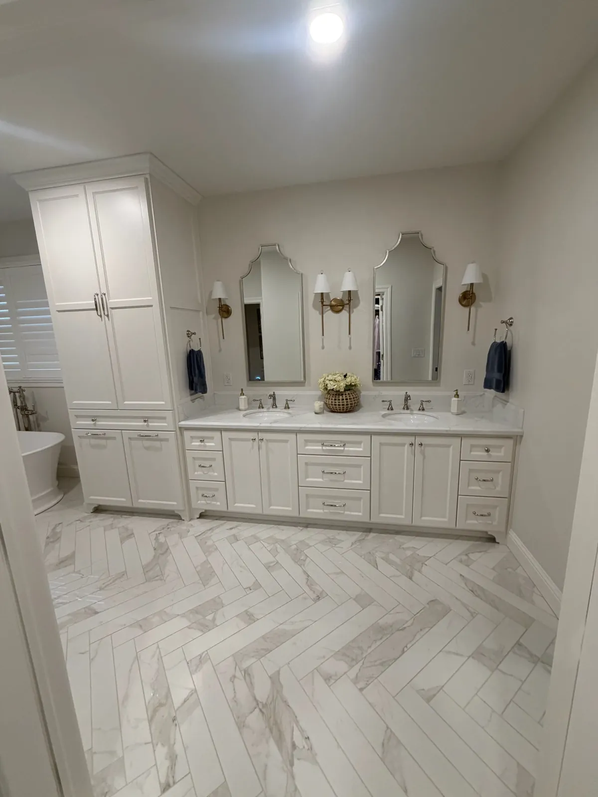 Primary bathroom with a white double vanity, linen storage tower and marble herringbone tile floor