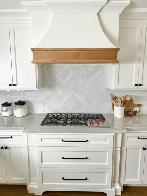 White kitchen with a plaster range hood, white oak mantel and herringbone marble backsplash