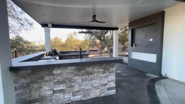 Covered outdoor kitchen with a stacked stone bar, granite counter and built-in grill under a beamed patio roof